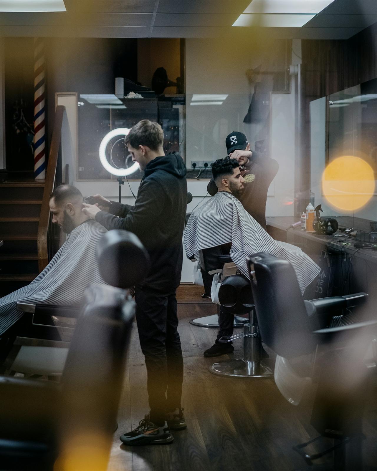 Two barbers at work in a dimly lit barbershop, warm amber light glowing behind the chairs
