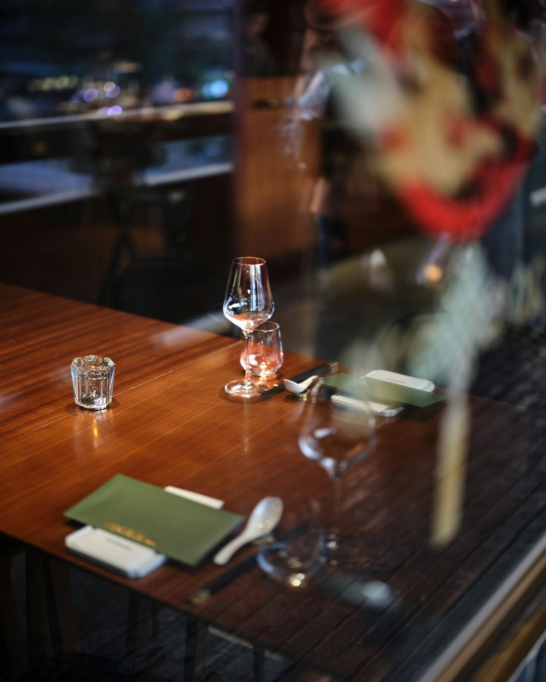 A dining table set for a meal beside a window, with a wine glass, menu and small vase