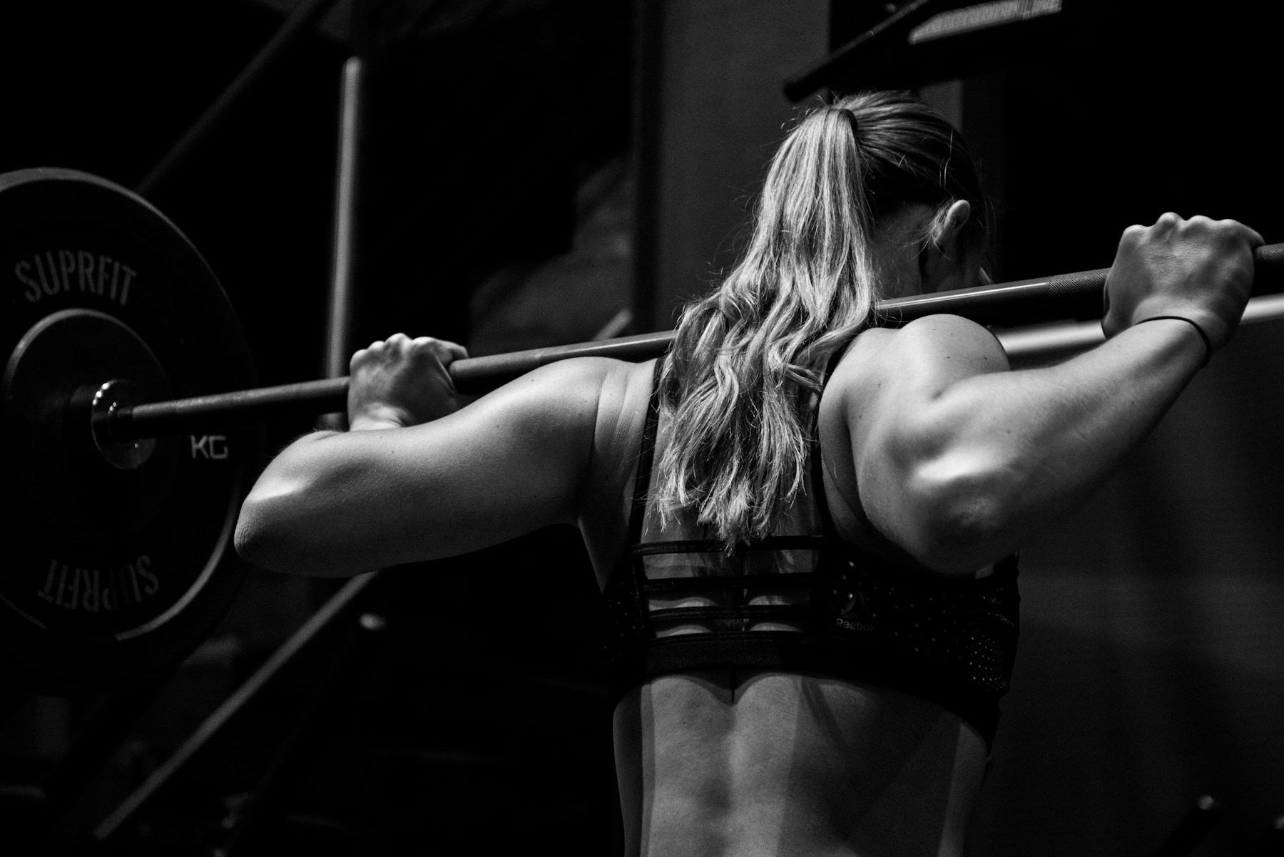 A woman athlete in black and white, seen from behind, gripping a loaded barbell across her shoulders