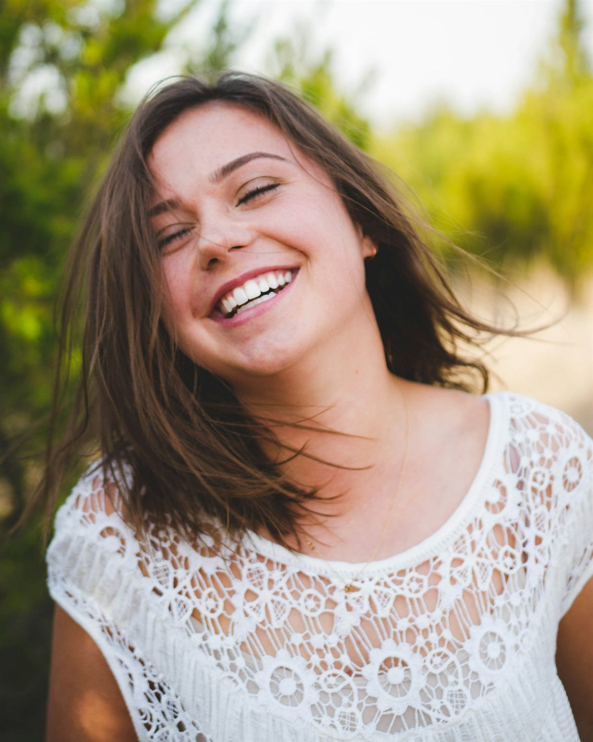 A young woman with windswept hair laughing in a white lace top, outdoors